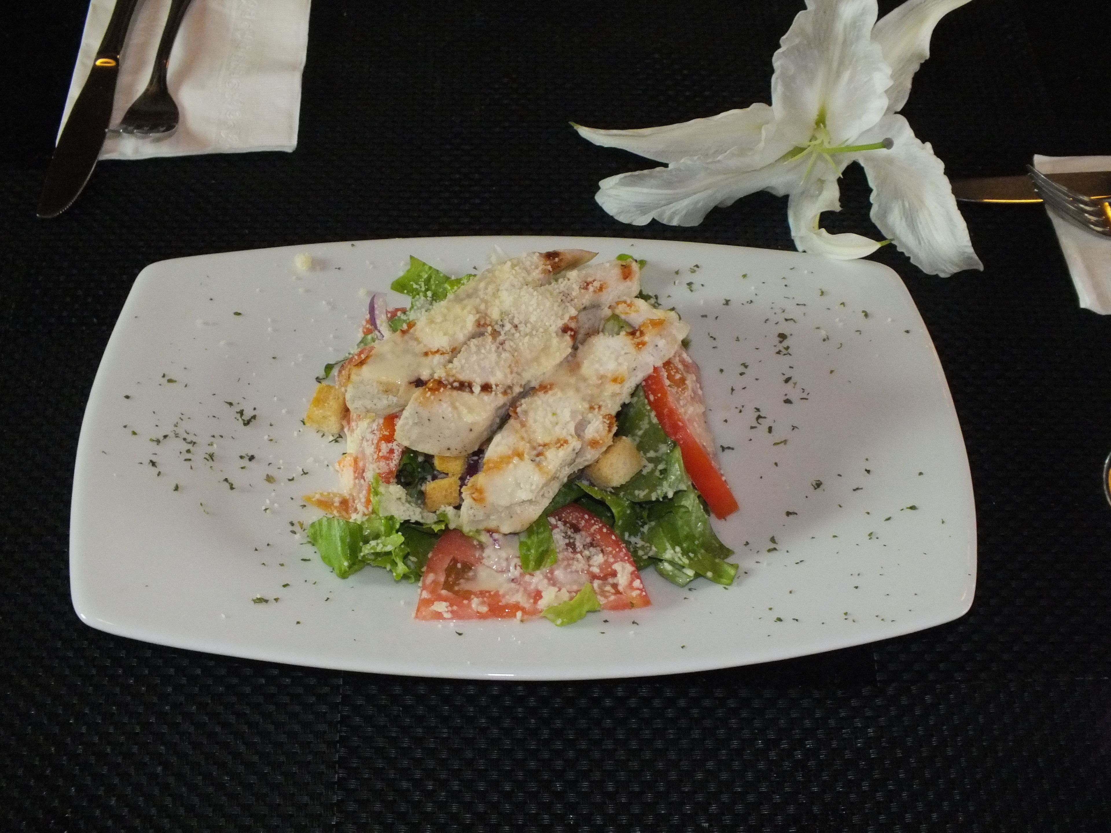 A white rectangular plate with a salad, including lettuce, tomatoes, and croutons, on a dark textured tablecloth.