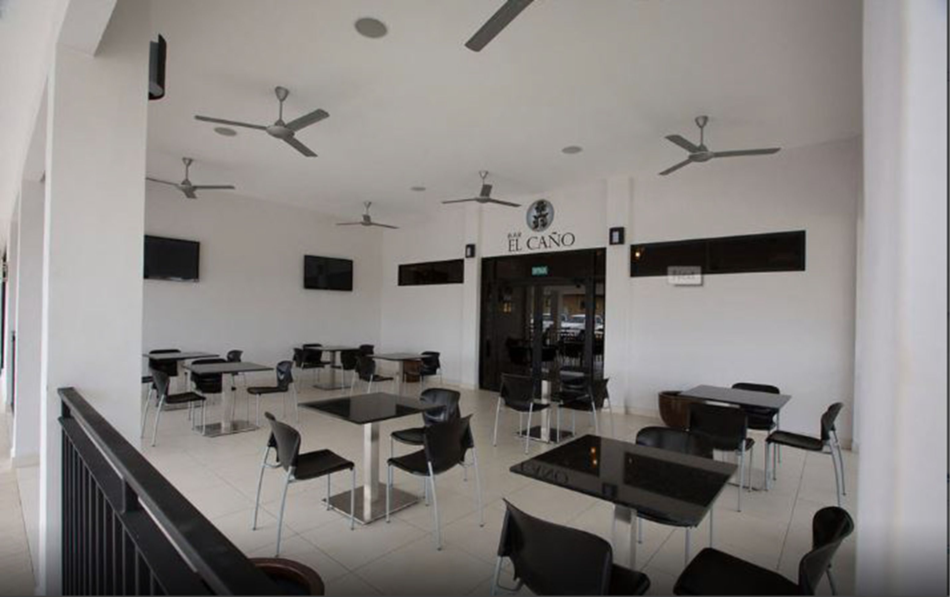 A modern restaurant interior with multiple tables and chairs, ceiling fans, and a visible entrance to another room labeled "EL CANO".
