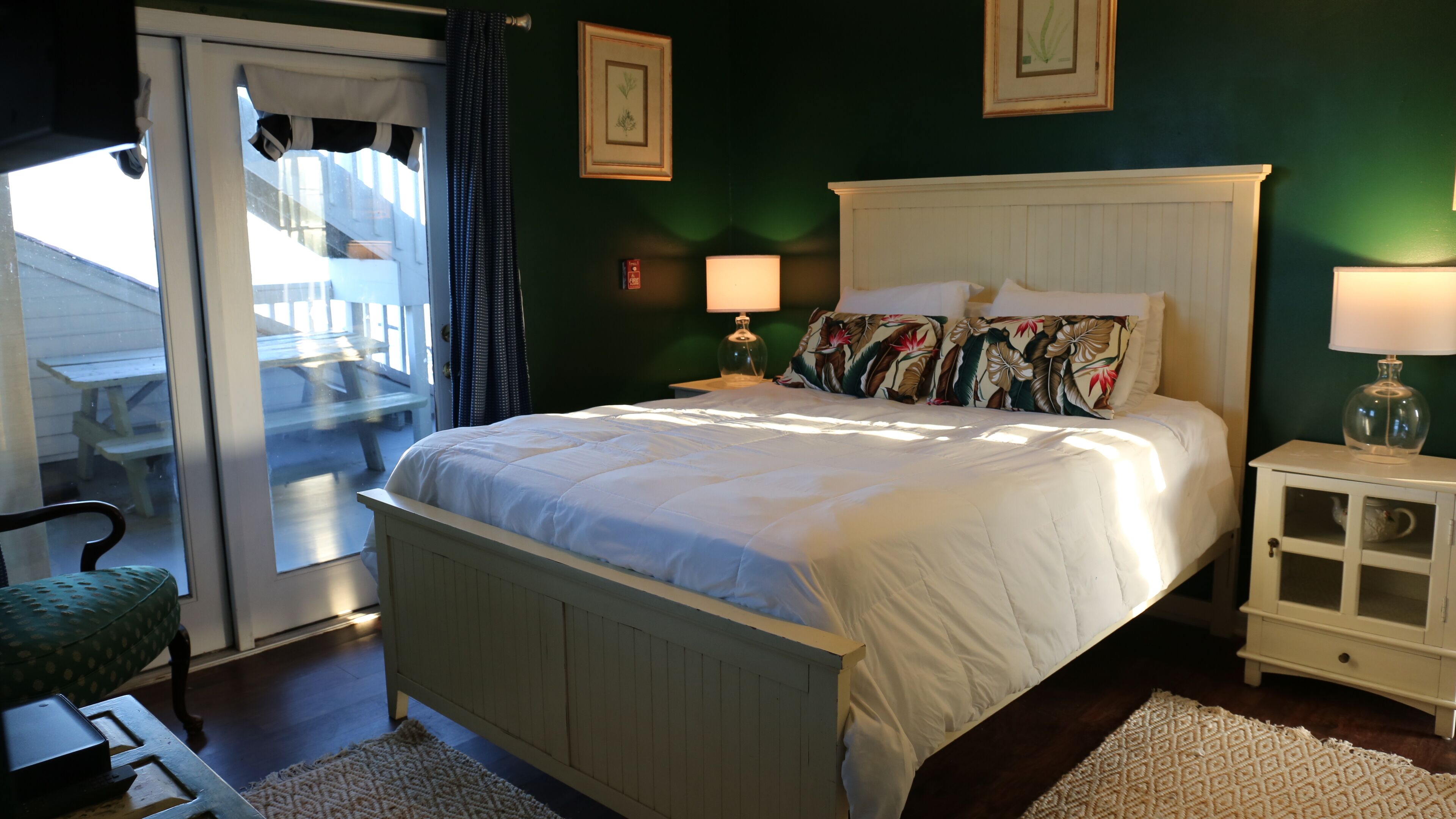 A bedroom with a large bed, two bedside lamps, a nightstand, a chair, and a view of an outdoor deck.