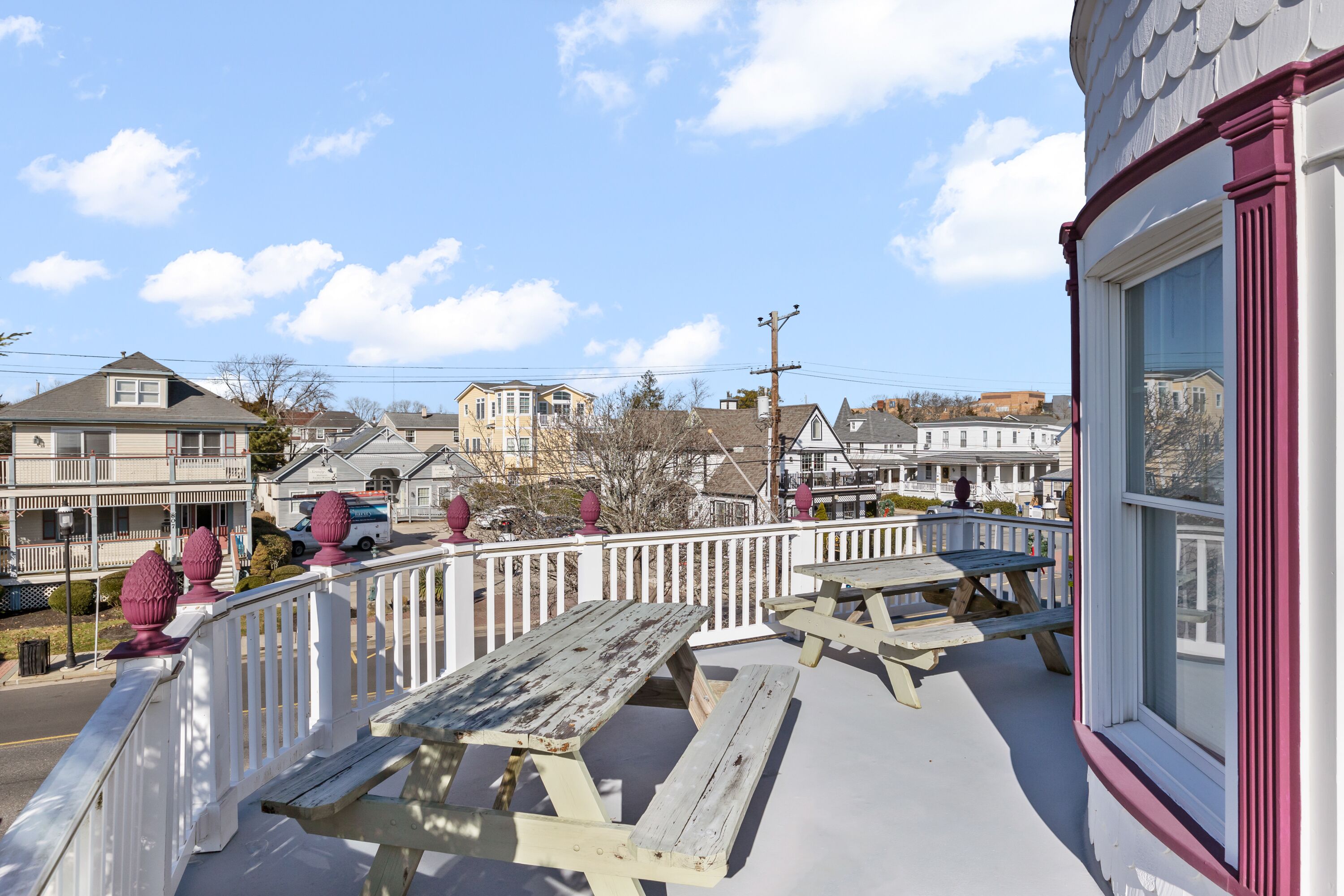 A balcony with picnic tables, a white railing, and a view of a residential area with houses and a clear sky.