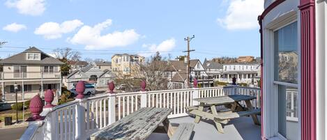 A balcony with picnic tables, a white railing, and a view of a residential area with houses and a clear sky.