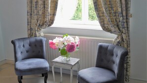 Two grey upholstered chairs with a small white table between them, a vase with pink and white flowers, and a window with curtains. - Chambres d'Hôtes Château de Courcelette (Lannoy)