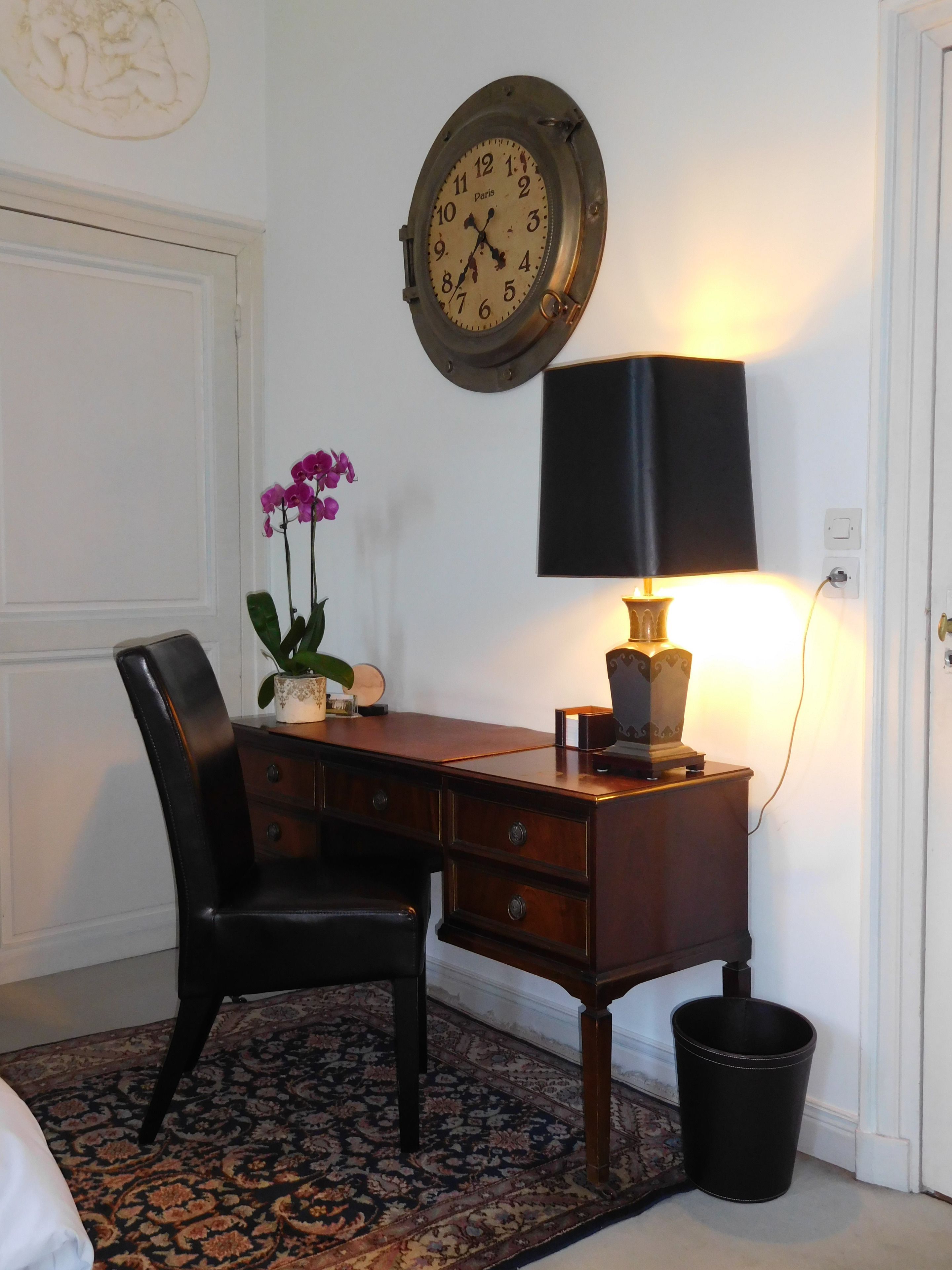 A room with a wooden desk, a black chair, a wall clock, a lamp, and a small trash bin.