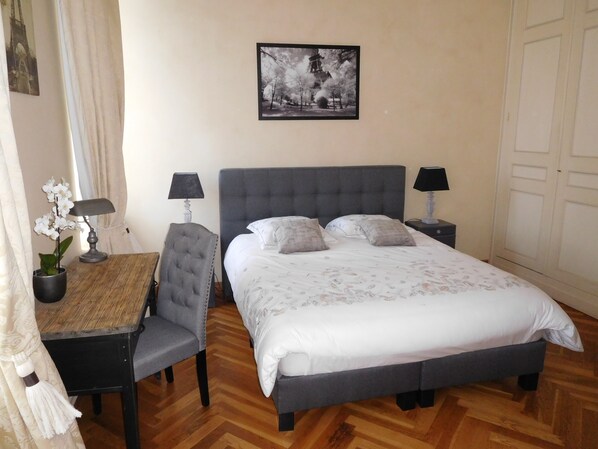 Un dormitorio ordenado con cama, dos mesitas de noche con lámparas, una silla, una pequeña mesa con lámpara y un cuadro colgado en la pared.