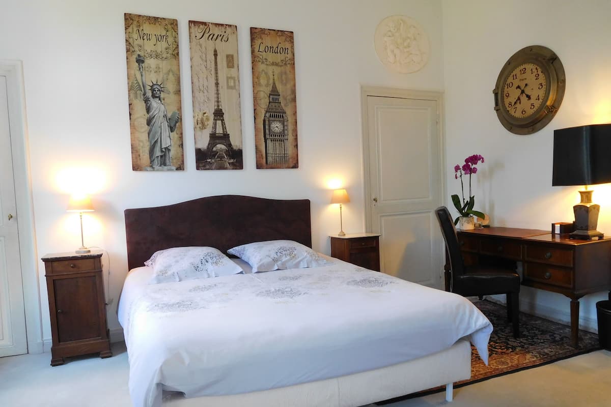 A bedroom with a bed, two bedside tables, a desk, a chair, a clock, and three wall decorations depicting famous landmarks.