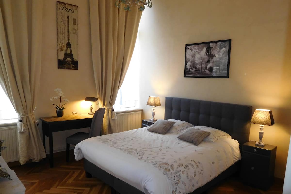 A bedroom with a large bed, two bedside tables with lamps, a desk with a chair, a radiator, and curtains.