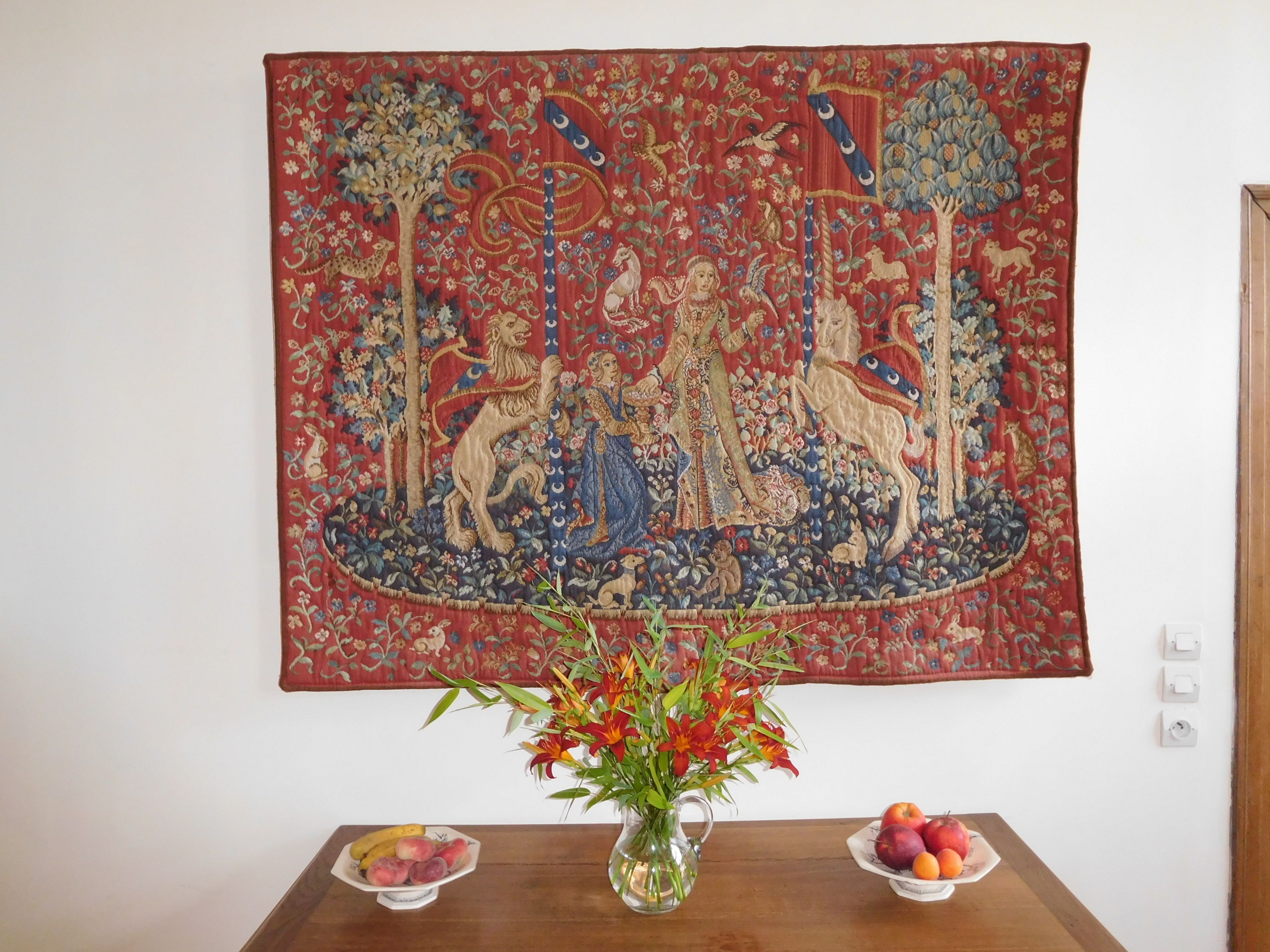 Un tapiz con una figura central y animales, un jarrón con flores, y dos cuencos de fruta sobre una mesa.
