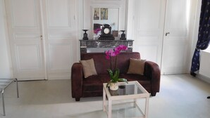 A living room with a dark brown sofa, two beige cushions, a glass coffee table with a potted orchid, and a fireplace with a clock and decorative items. - Chambres d'Hôtes Château de Courcelette (Lannoy)