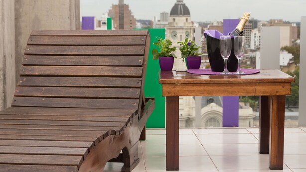 Una silla lounge de madera y una mesita con una botella de champán, dos jarrones morados y dos plantas en macetas en un balcón con vista a la ciudad.