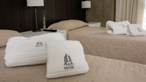 Habitación de hotel con dos camas, cada una con cubrecama y almohadas color beige. Dos toallas blancas con un logotipo, colocadas sobre las camas. Una lámpara y una pequeña planta están sobre la mesita de noche al lado de las camas.