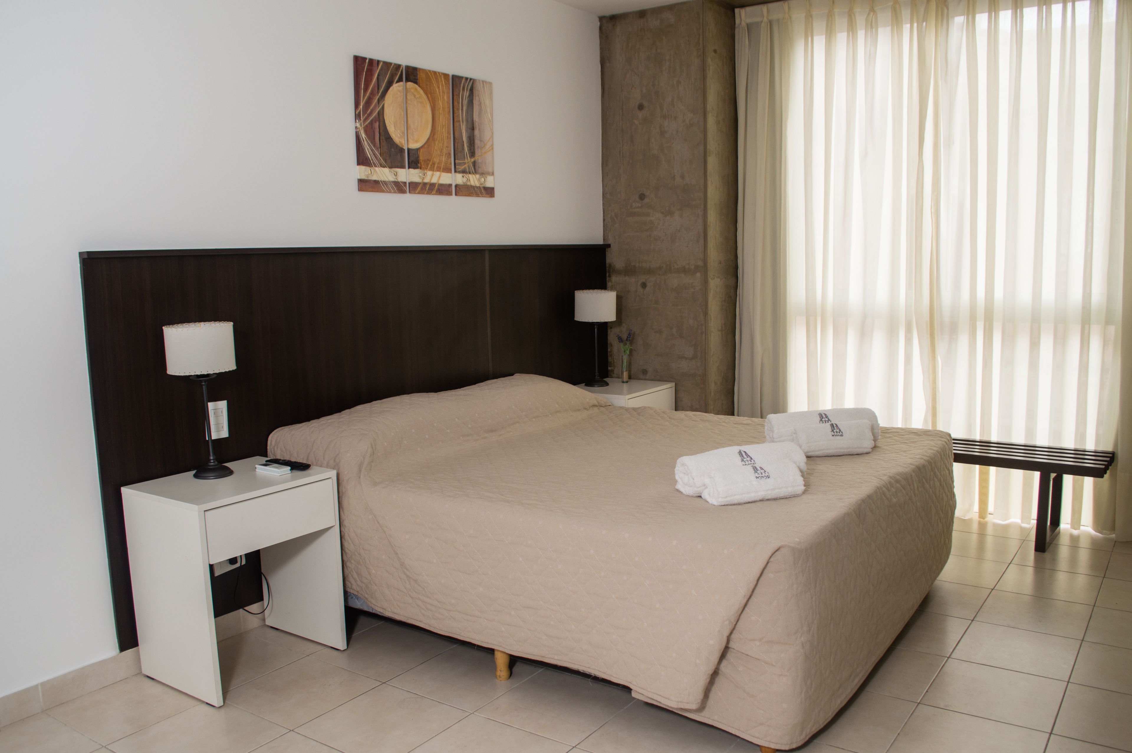 Een netjes opgemaakt bed met een beige sprei, twee nachtkastjes met lampen, een kunstwerk aan de muur en een raam met dunne gordijnen.