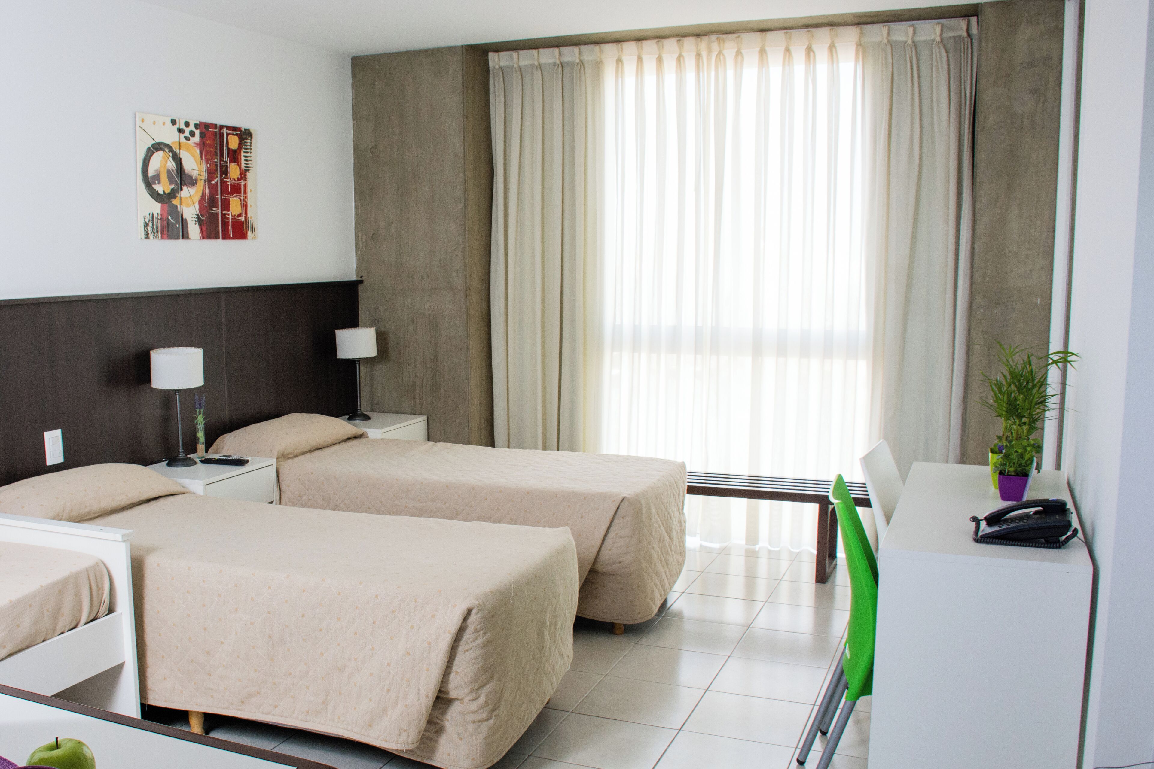 A hotel room with two beds, a desk, a chair, a lamp, a plant, and a window with curtains.