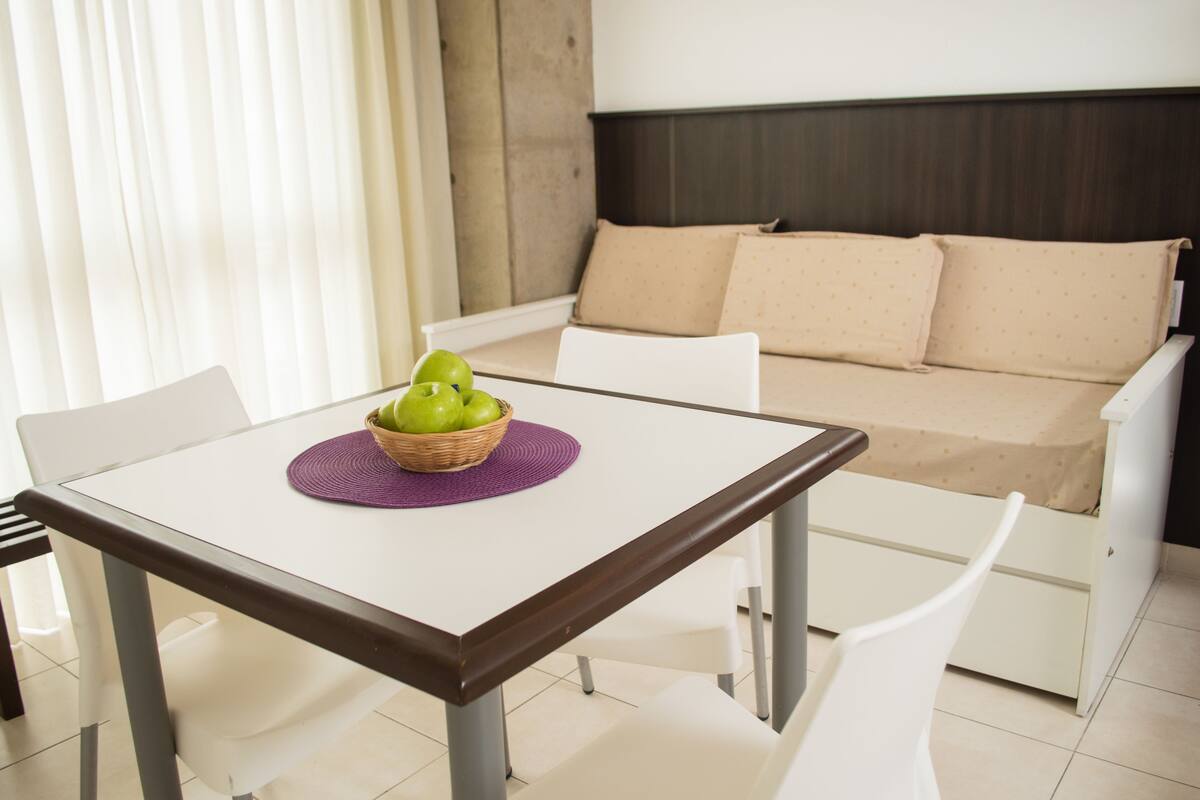Una habitación moderna con un sofá blanco, una mesa de comedor con camino de mesa morado y una cesta de manzanas verdes.