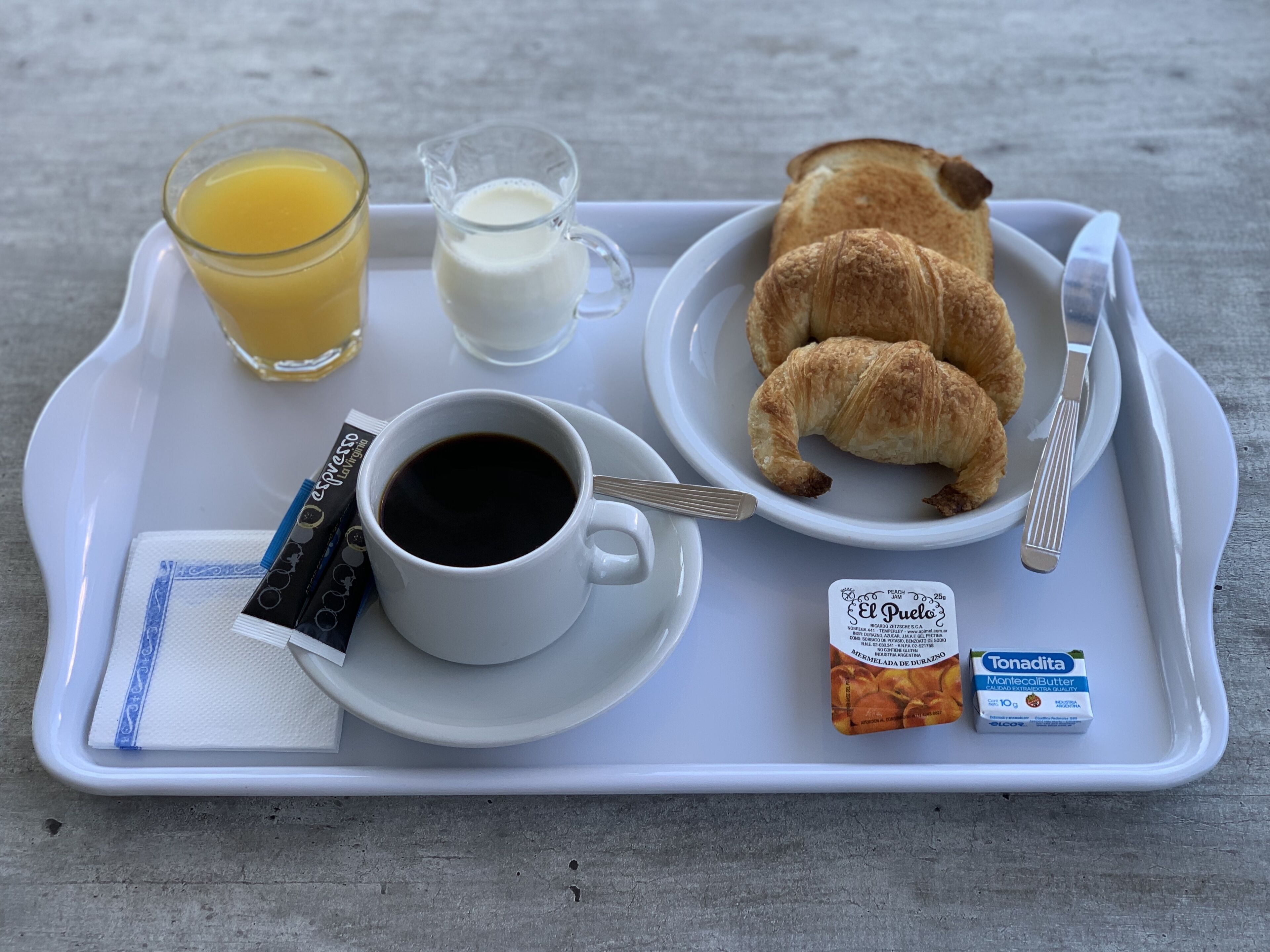A breakfast tray with croissants, a cup of coffee, a glass of orange juice, a jug of milk, a packet of instant coffee, and a carton of milk.