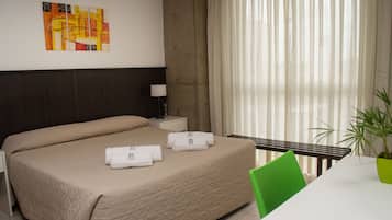 Una camera d'albergo con un letto, un comodino, una lampada, una sedia, un tavolo con un telefono e una pianta.
