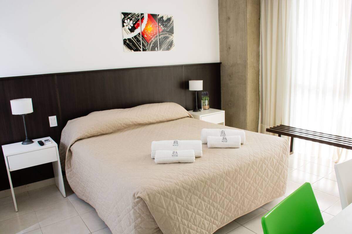 Una habitación de hotel con una cama, una mesita de noche con lámpara, una silla y un cuadro en la pared.