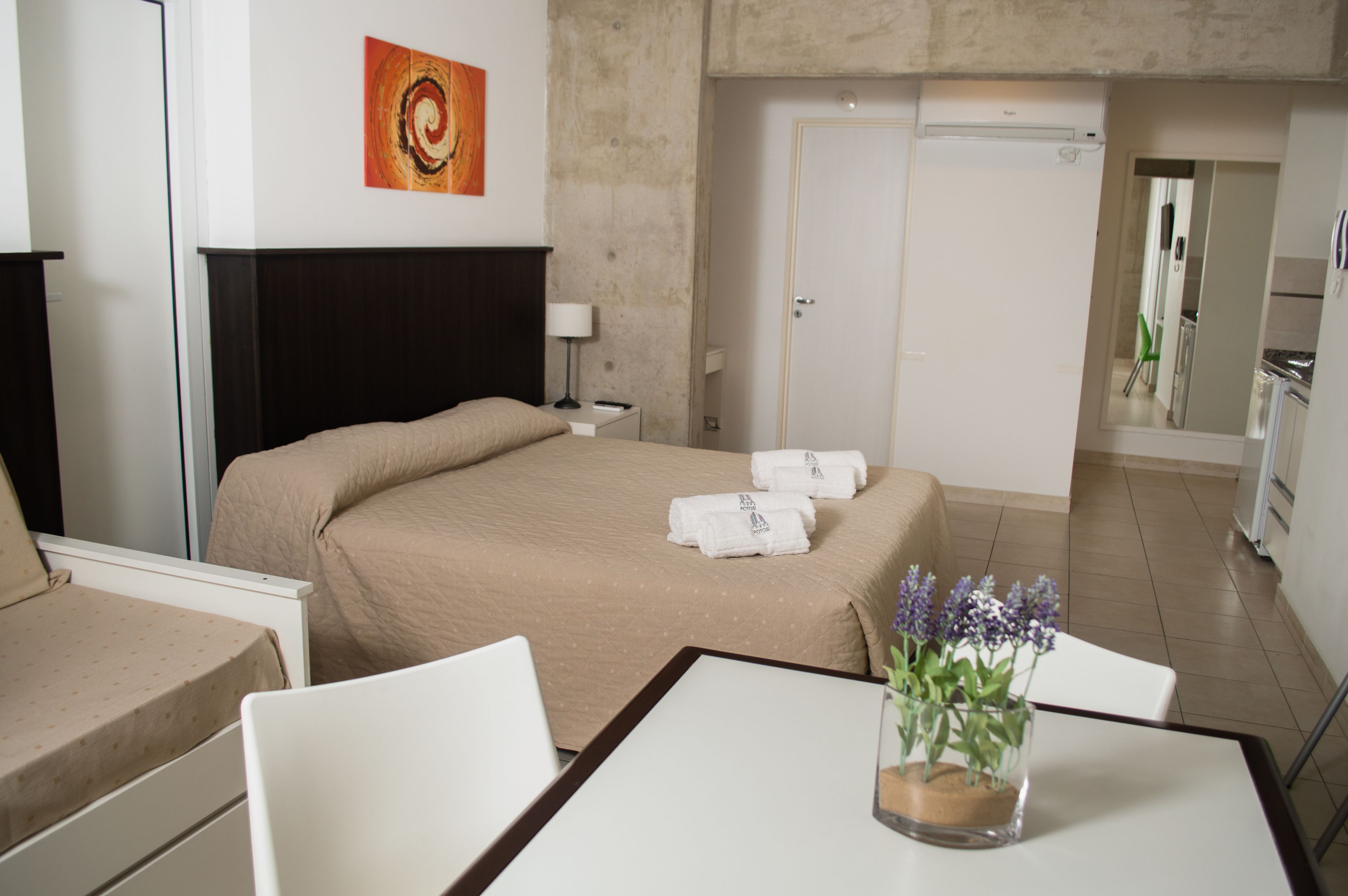 A hotel room with a bed, a desk, a chair, a lamp, a vase with flowers, a painting on the wall, and a door.