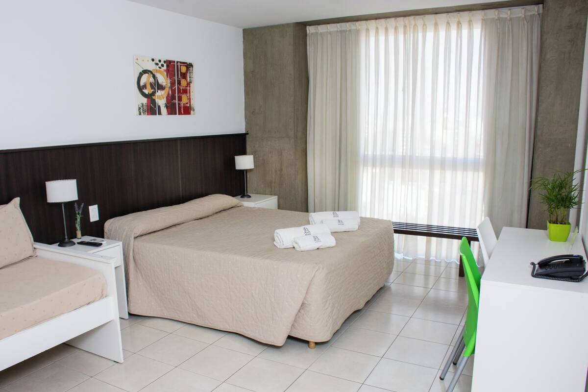 Habitación de hotel con una cama grande, un escritorio con silla, una planta en maceta y una ventana con cortinas.