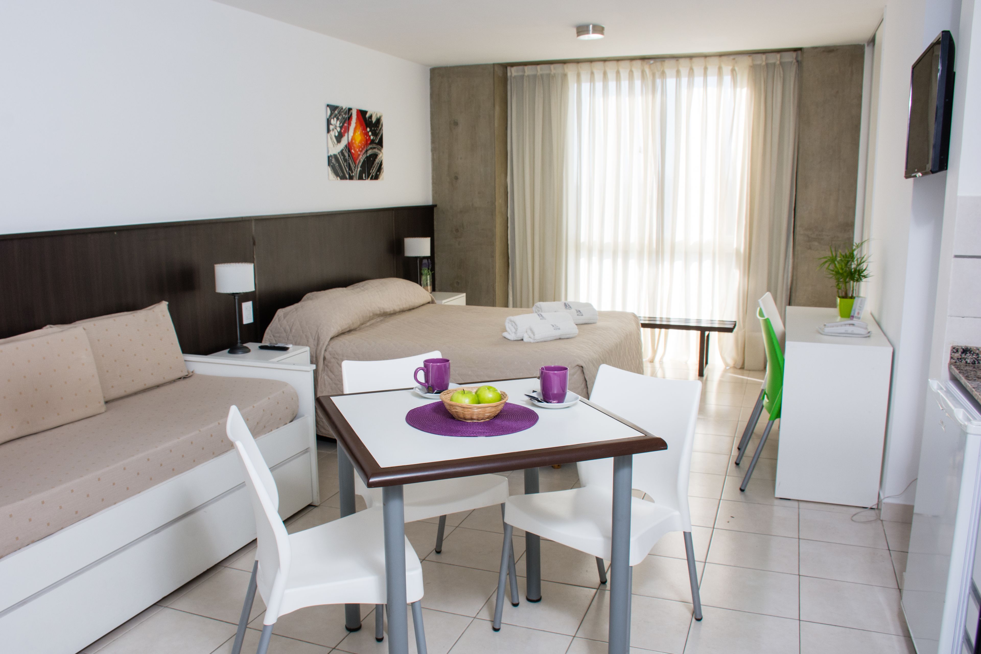 A hotel room with a bed, a sofa, a dining table with chairs, a small kitchenette, and a TV.