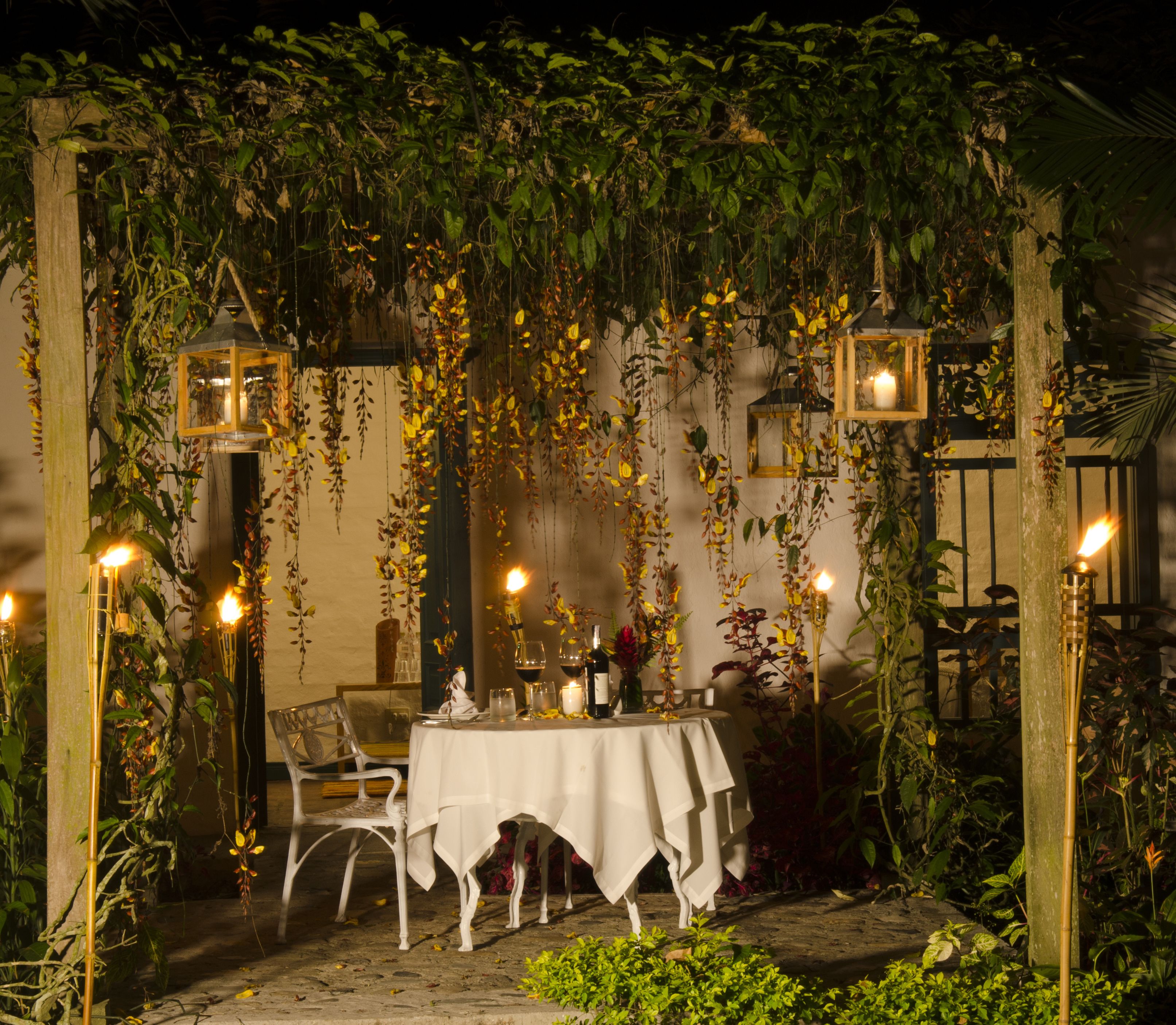 Un área de comedor al aire libre con una mesa preparada para dos personas, rodeada de follaje y faroles colgantes.
