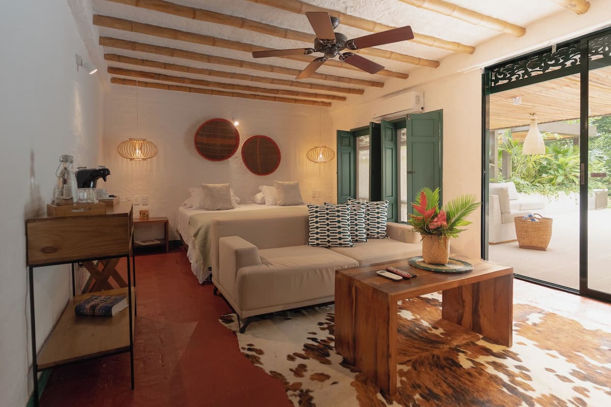 Une pièce confortable avec un lit, un canapé, une table basse et un coin repas. On y trouve un ventilateur de plafond, une lampe fixée au mur et une fenêtre donnant sur un paysage verdoyant.