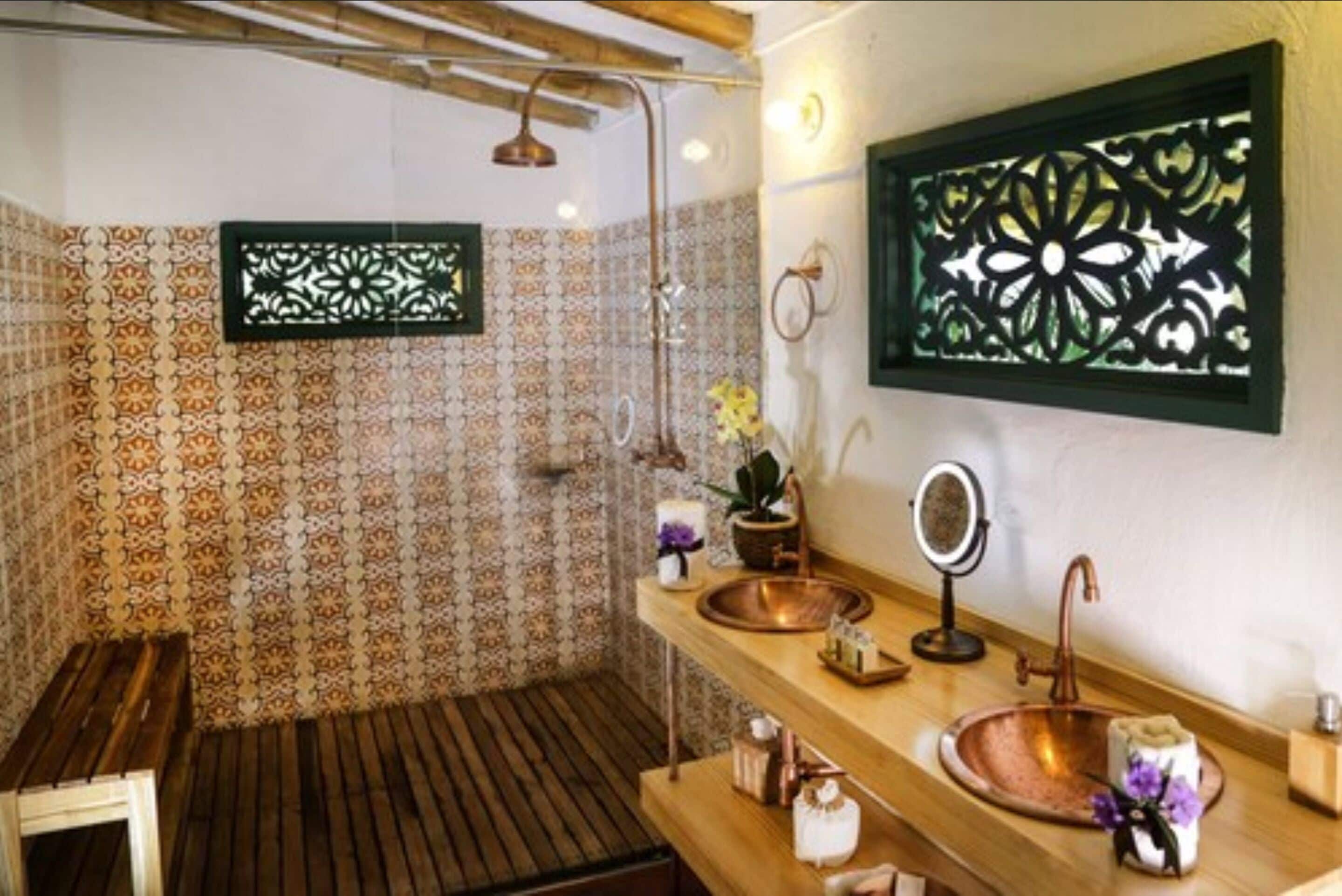 Un baño con baldosas estampadas, un banco de madera, un lavamanos de cobre, un espejo y paneles decorativos en la pared.