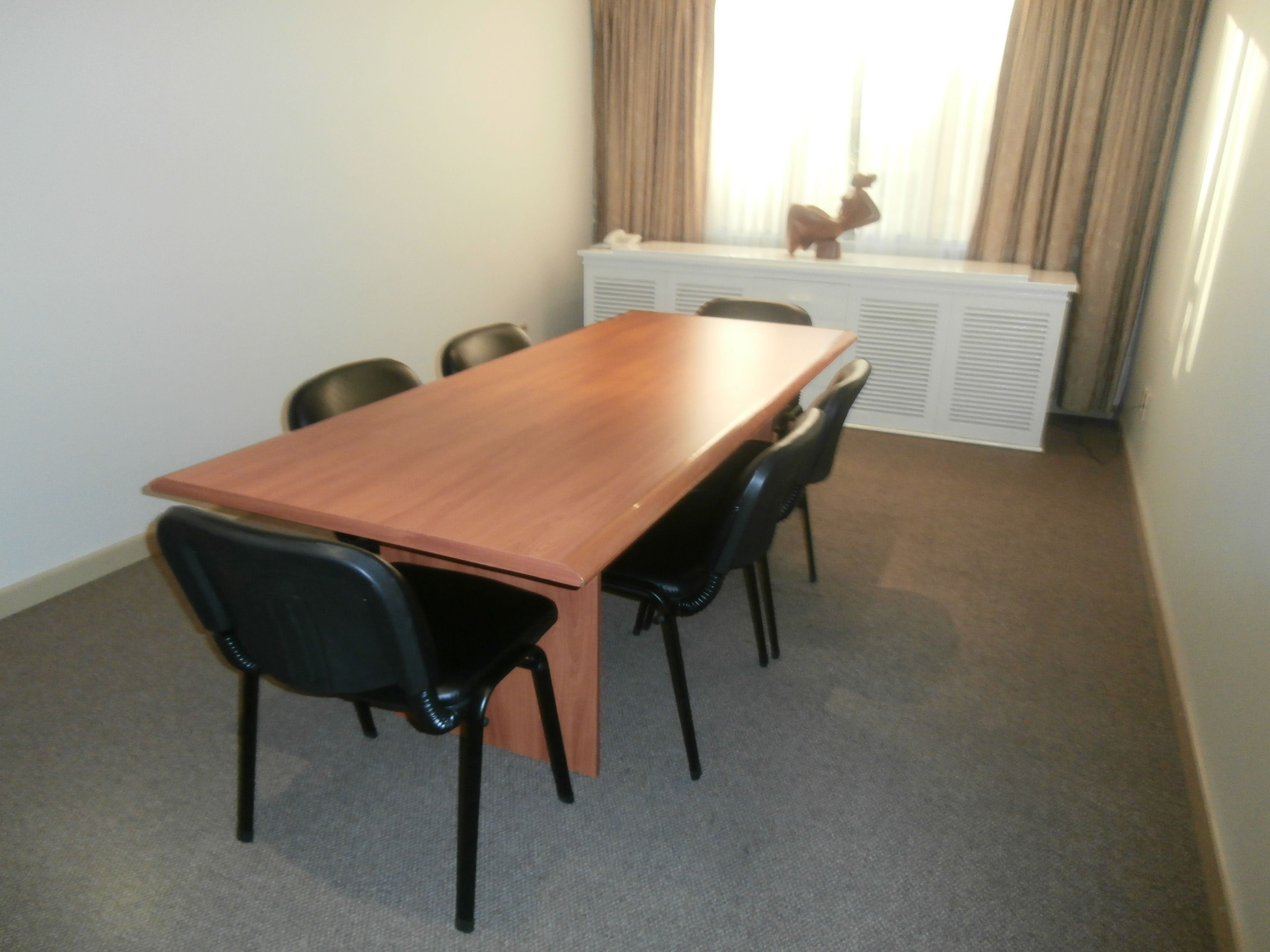 Una sala de conferencias con una mesa de madera, seis sillas negras, un radiador blanco y un ventanal con cortinas.