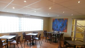 Una habitación amplia con varias mesas y sillas, ventanas grandes con persianas y un cuadro abstracto moderno en la pared.
