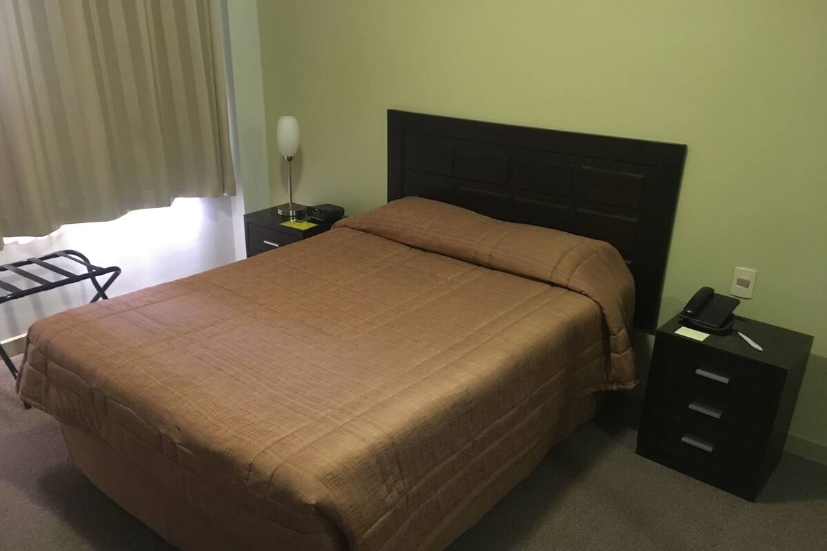 A neatly made bed with a brown blanket, a bedside table with a lamp, and a phone.
