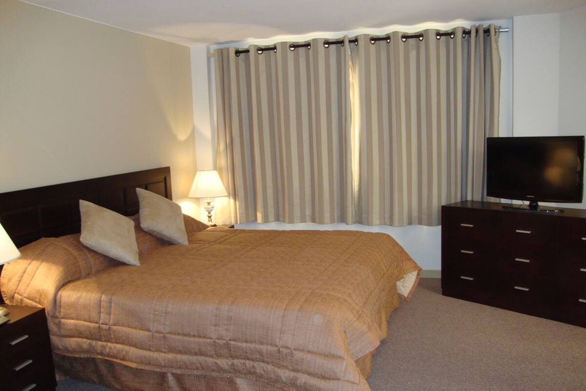 A hotel room with a large bed, a TV on a dresser, a lamp, and a window with curtains.