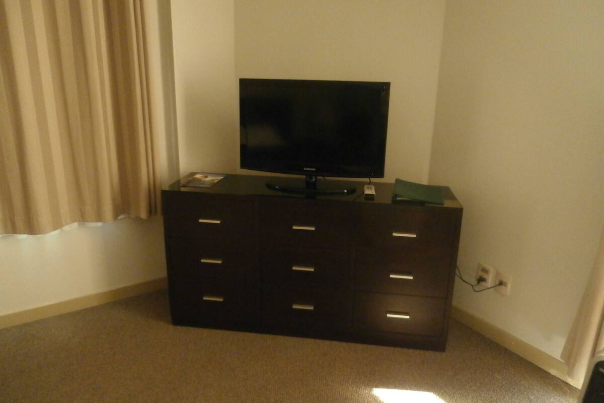 A dark wooden dresser with a flat-screen TV on top, a remote control, and a small electronic device on the side.