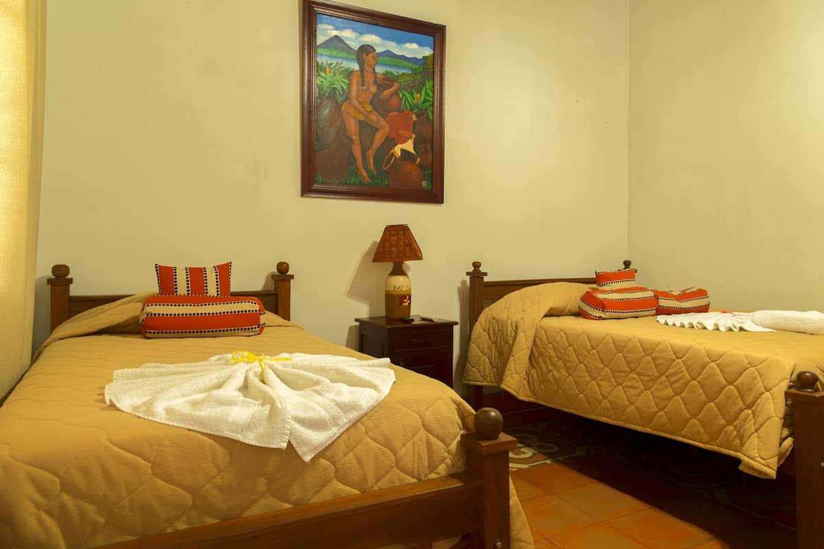 A hotel room with two beds, a painting on the wall, a nightstand with a lamp, and a window with curtains.
