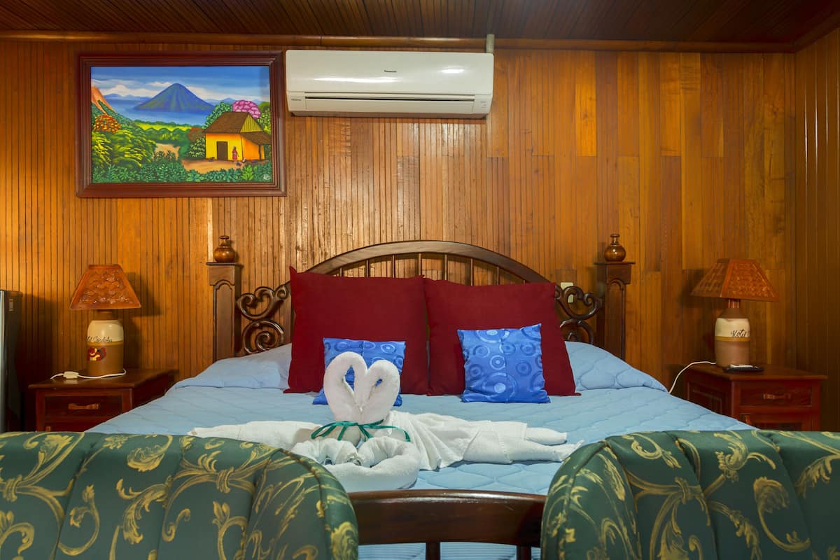 A hotel room with a wooden paneled wall, a bed with a blue quilt, red and blue pillows, and a painting of a landscape.