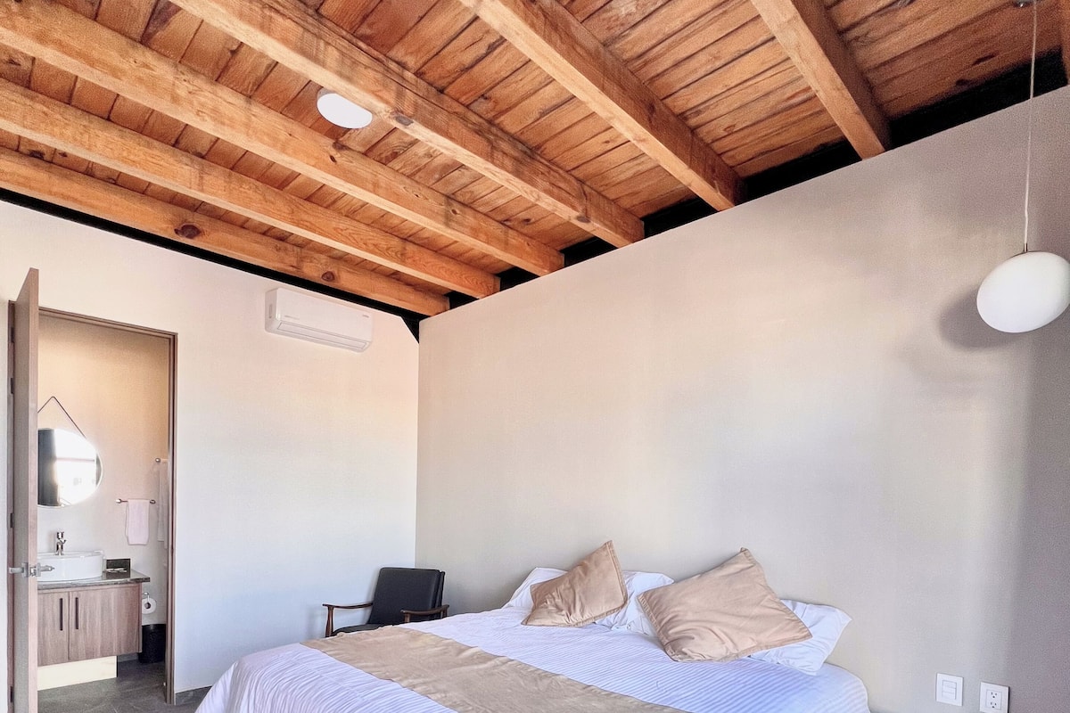 A bedroom with a large bed, a wooden ceiling, a small bedside table, and a bathroom visible through an open door.