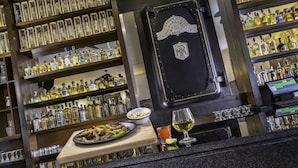 Un bar avec des étagères remplies de bouteilles d’alcool, un comptoir en bois sur lequel se trouve une assiette de nourriture et un verre de vin, et une caisse enregistreuse.