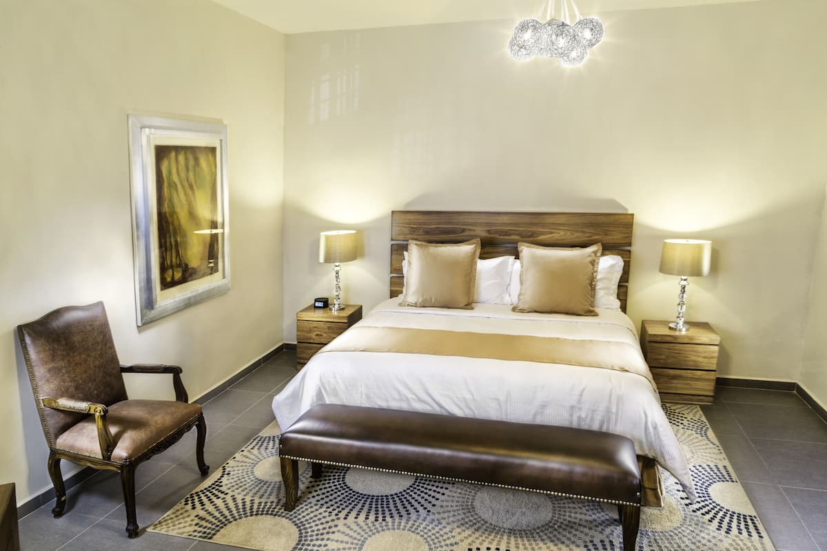 A hotel room with a large bed, two bedside tables, a chair, a bench, a patterned rug, and a chandelier.