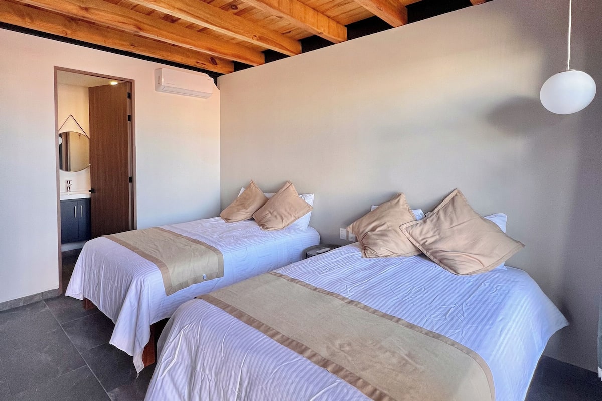 A room with two beds, wooden ceiling beams, a hanging light fixture, and a door leading to a bathroom.