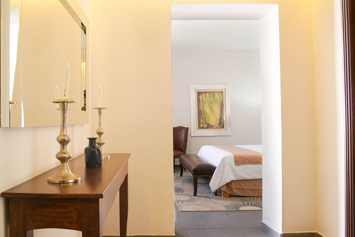 A hotel room with a wooden desk, a mirror, a bed with a white bedspread, a brown leather chair, and a painting on the wall.