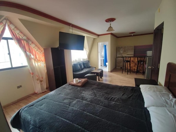 Une chambre avec un grand lit, un téléviseur fixé au mur, un canapé et un coin repas avec des chaises.