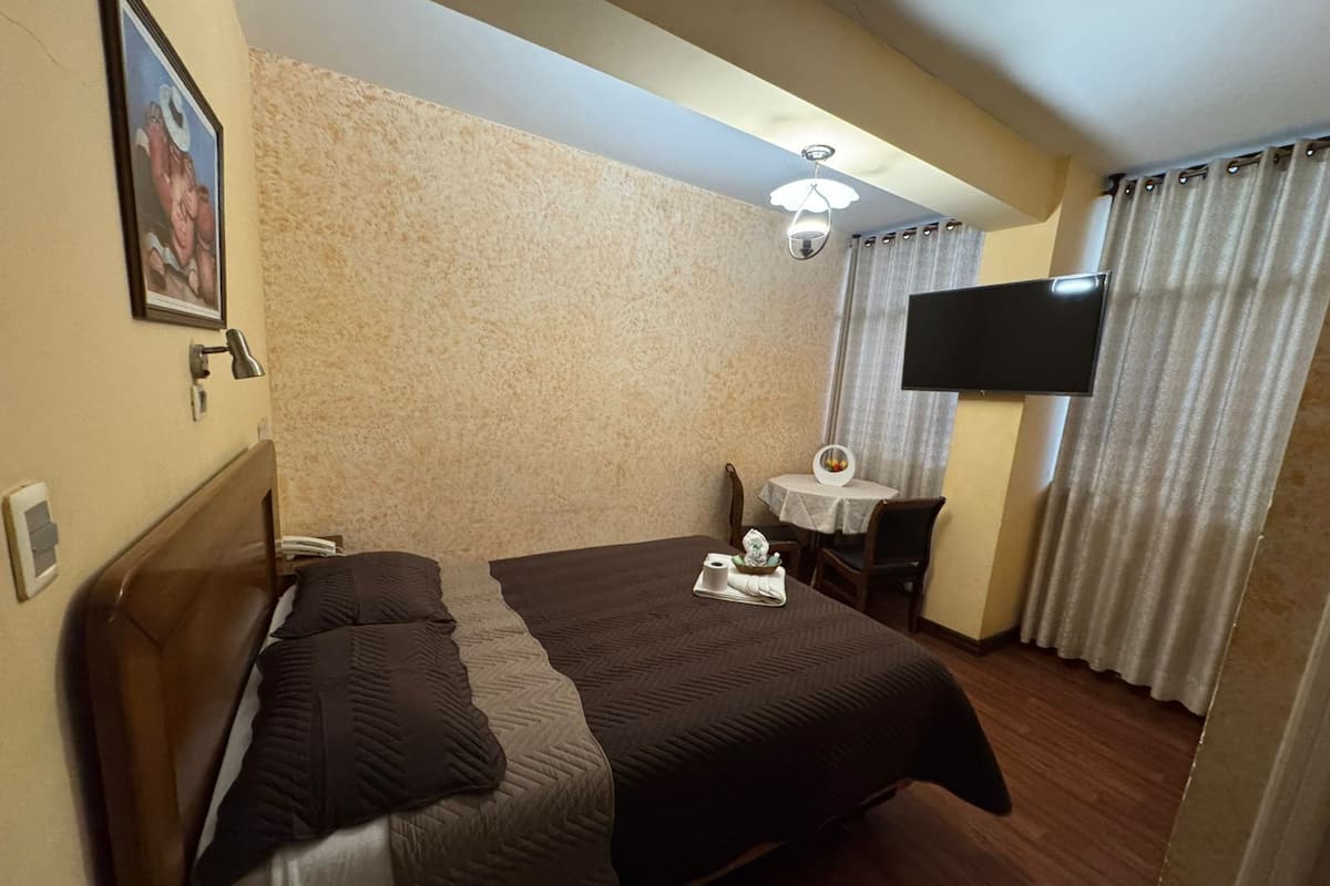 A hotel room with a bed, a desk, a chair, a TV, and a small table with a decorative item.