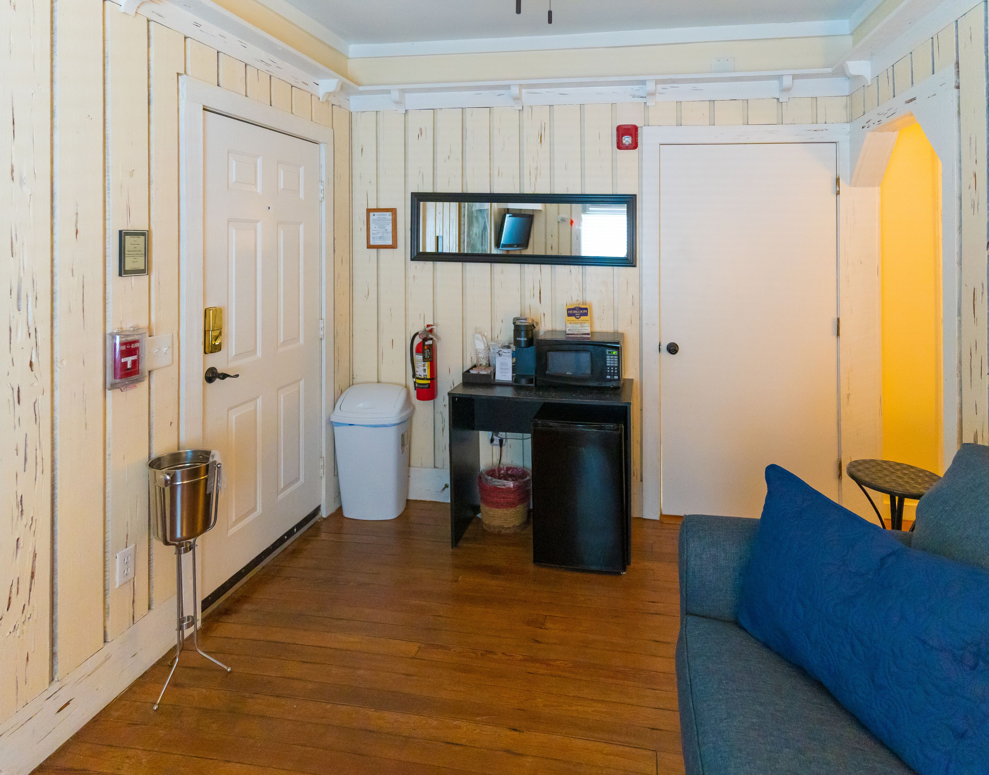 A compact room with a wooden floor, a blue couch, a small table, a microwave, a trash bin, and a fire extinguisher.