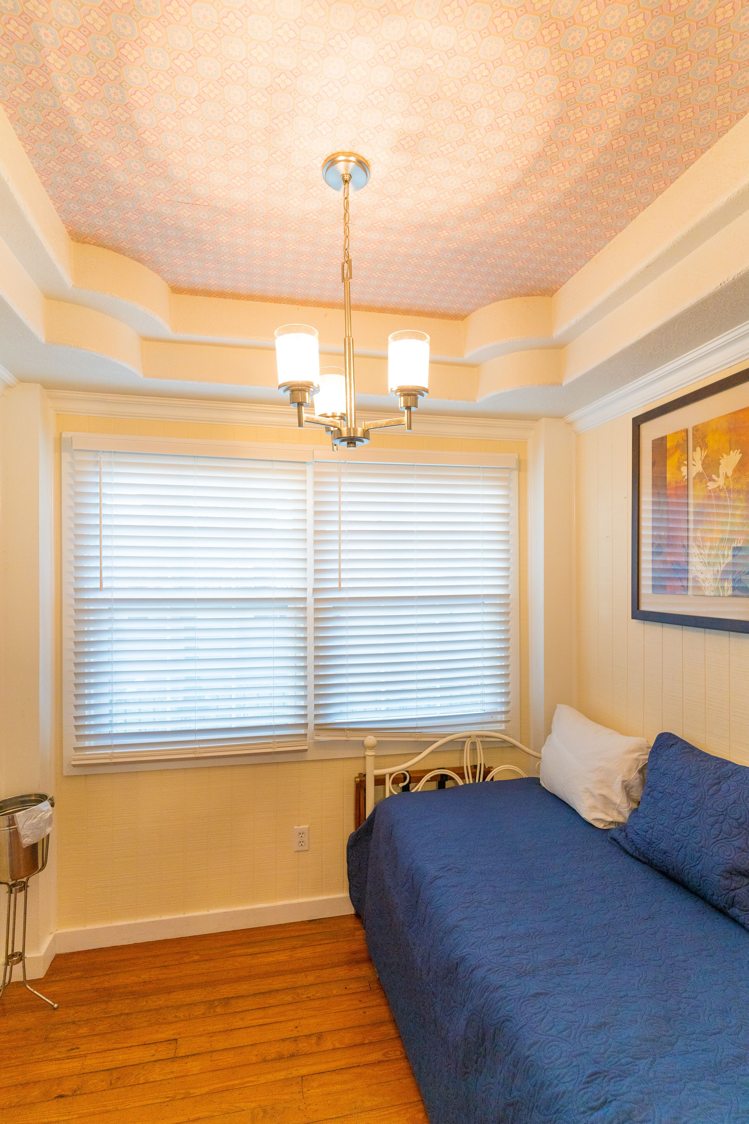 Une chambre à coucher avec un lit, une fenêtre avec des stores, un ventilateur de plafond et un luminaire fixé au mur.
