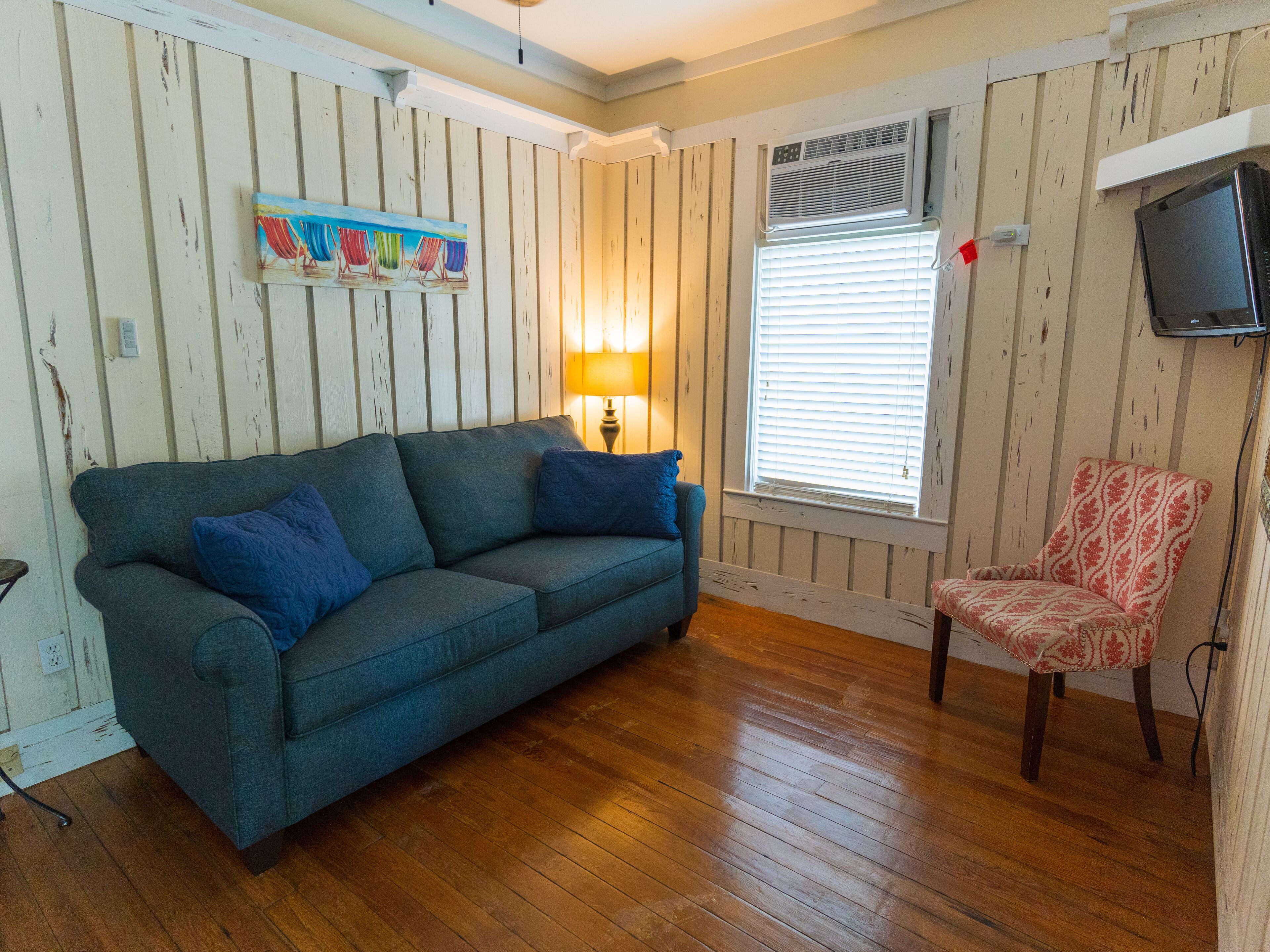 Una habitación con un sofá azul, un sillón estampado, un cuadro, una lámpara y un televisor en la pared.