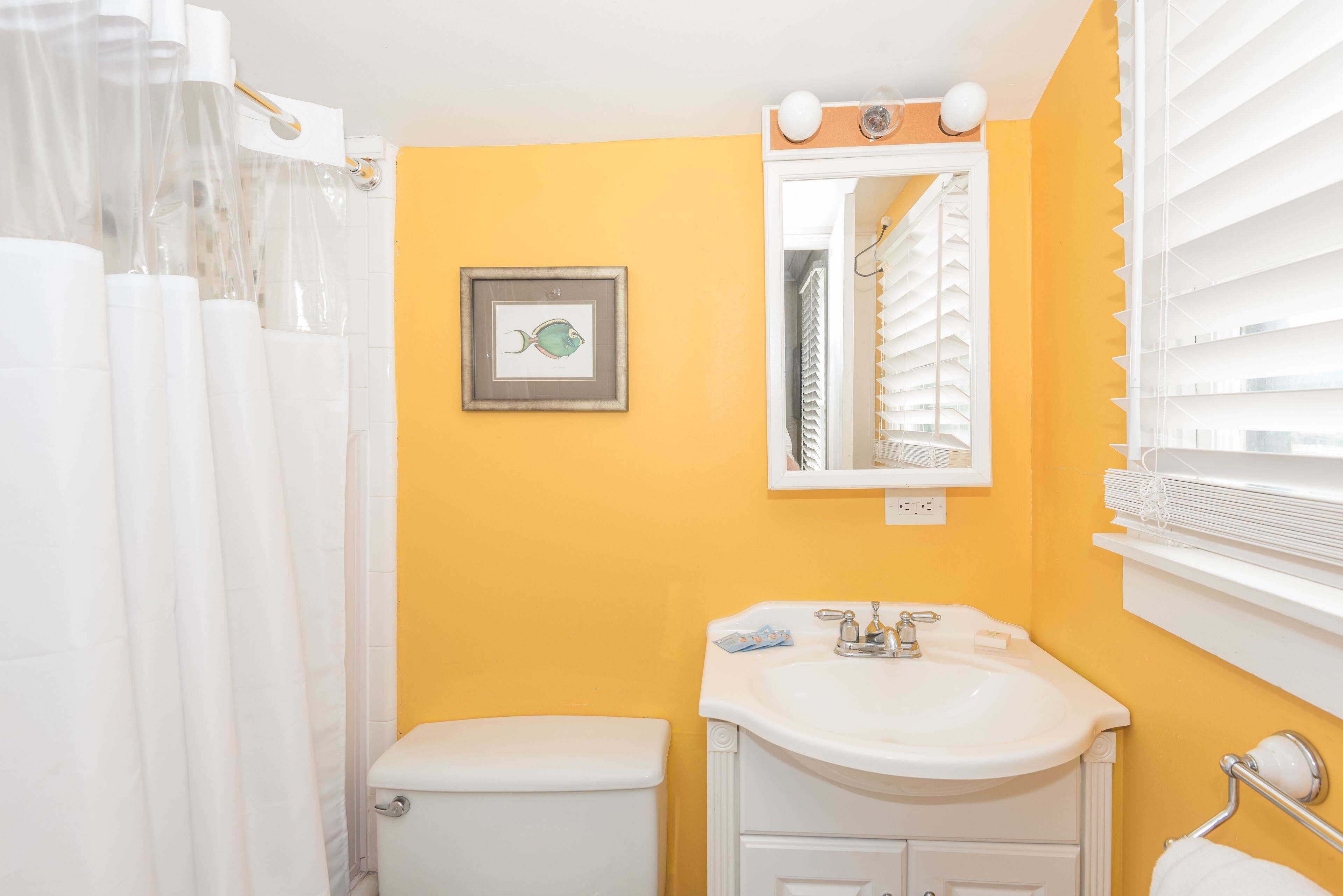 A bathroom with a yellow wall, a white sink, a framed picture, and a window with blinds.