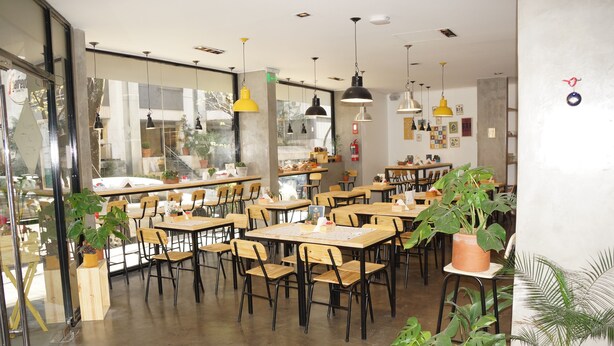Un café con mesas y sillas de madera, ventanas amplias y lámparas colgantes.