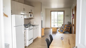 A compact kitchen with white appliances, a dining area with a wooden table and chairs, and a large window providing natural light.