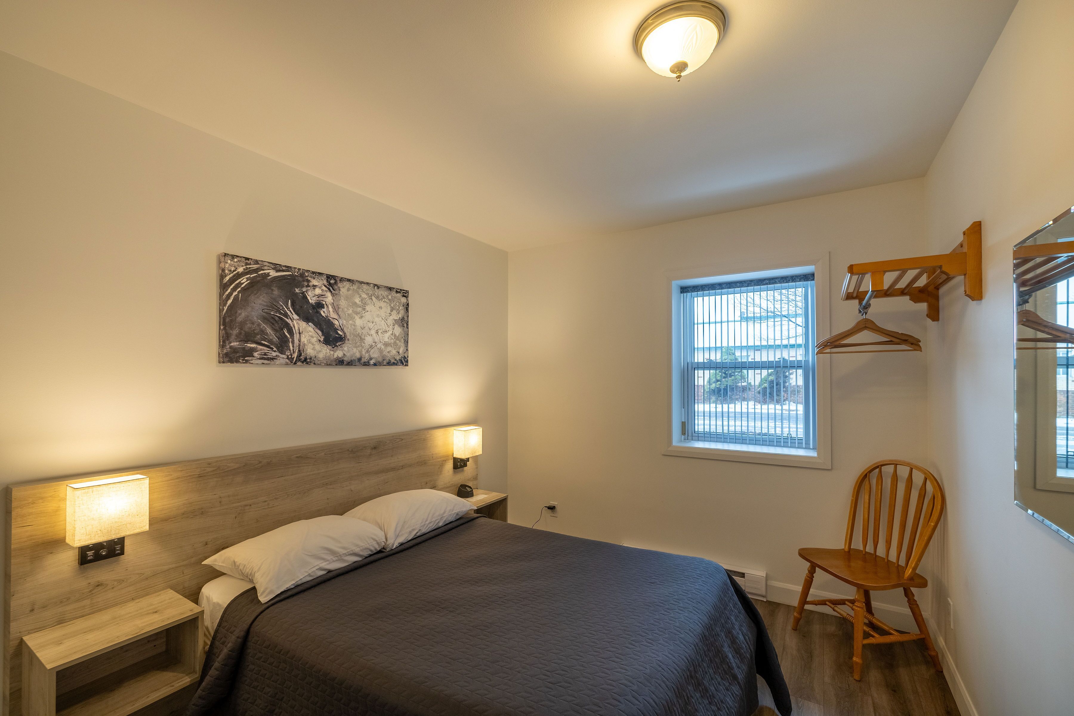 A bedroom with a bed, bedside lamps, a wooden headboard, a wall-mounted artwork, a window with blinds, and a wooden chair.