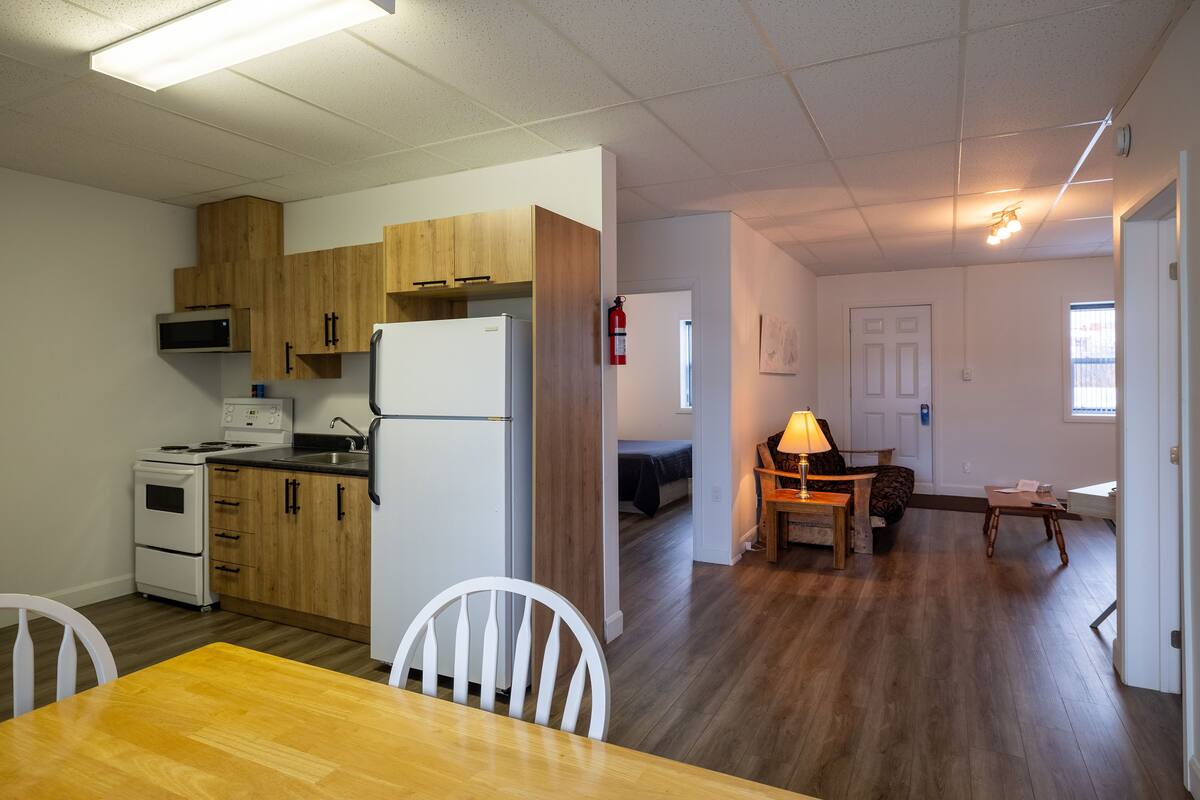 A compact kitchen with wooden cabinets, a white refrigerator, and a microwave. There is a dining table with chairs and a small wooden table with a lamp. The room has hardwood flooring and a door leading to another room.
