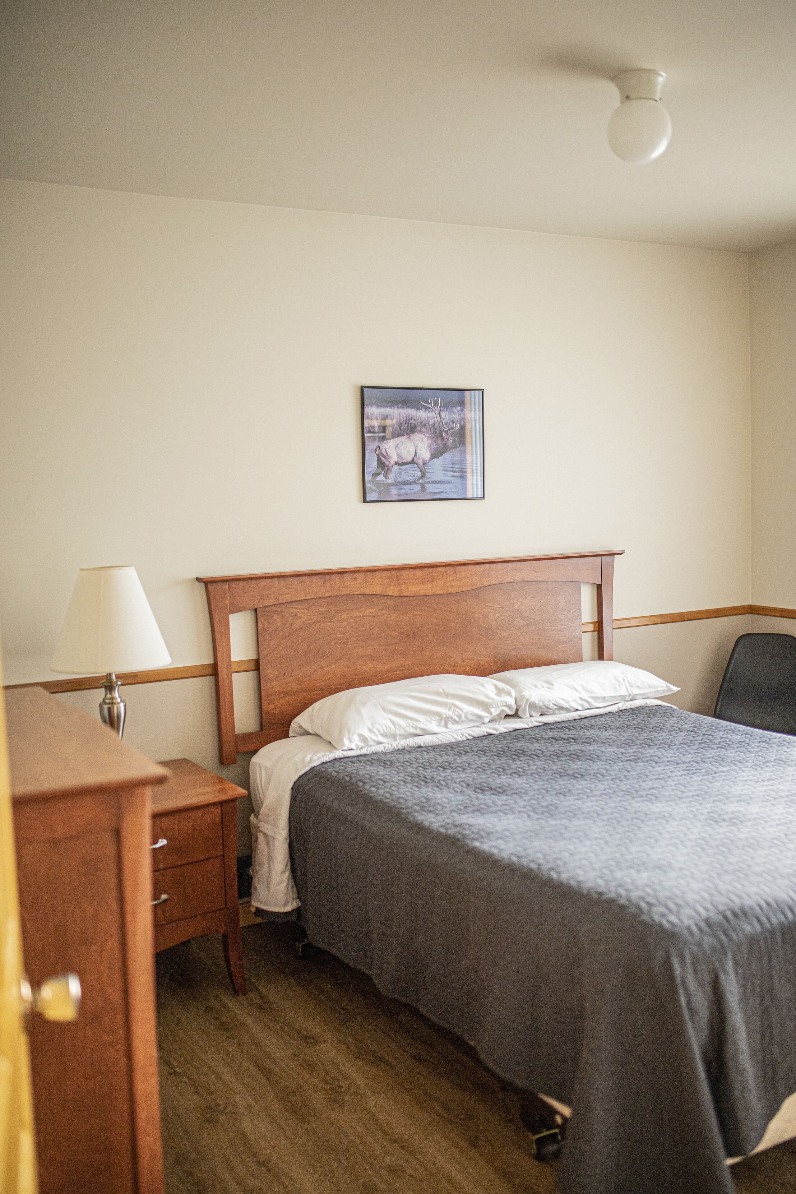 Une chambre à coucher avec un lit en bois, une table de chevet, une lampe, une chaise et un tableau encadré accroché au mur.
