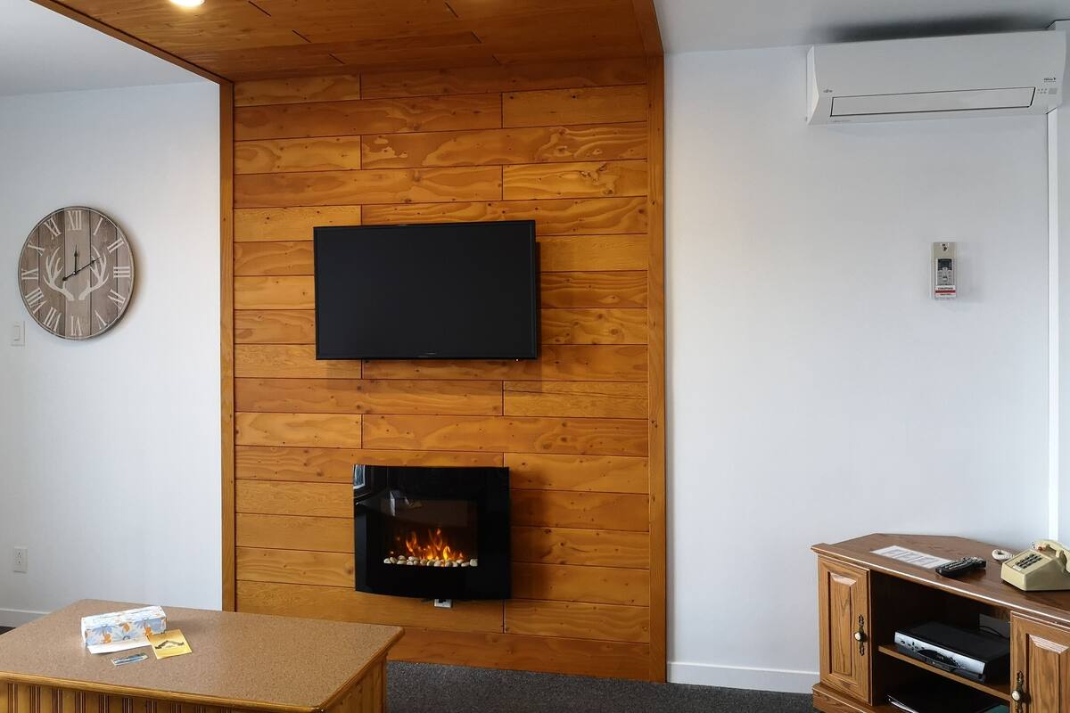 A modern living room with a wooden wall panel, a flat-screen TV, a fireplace, a clock, and a wooden cabinet with a telephone and remote controls.